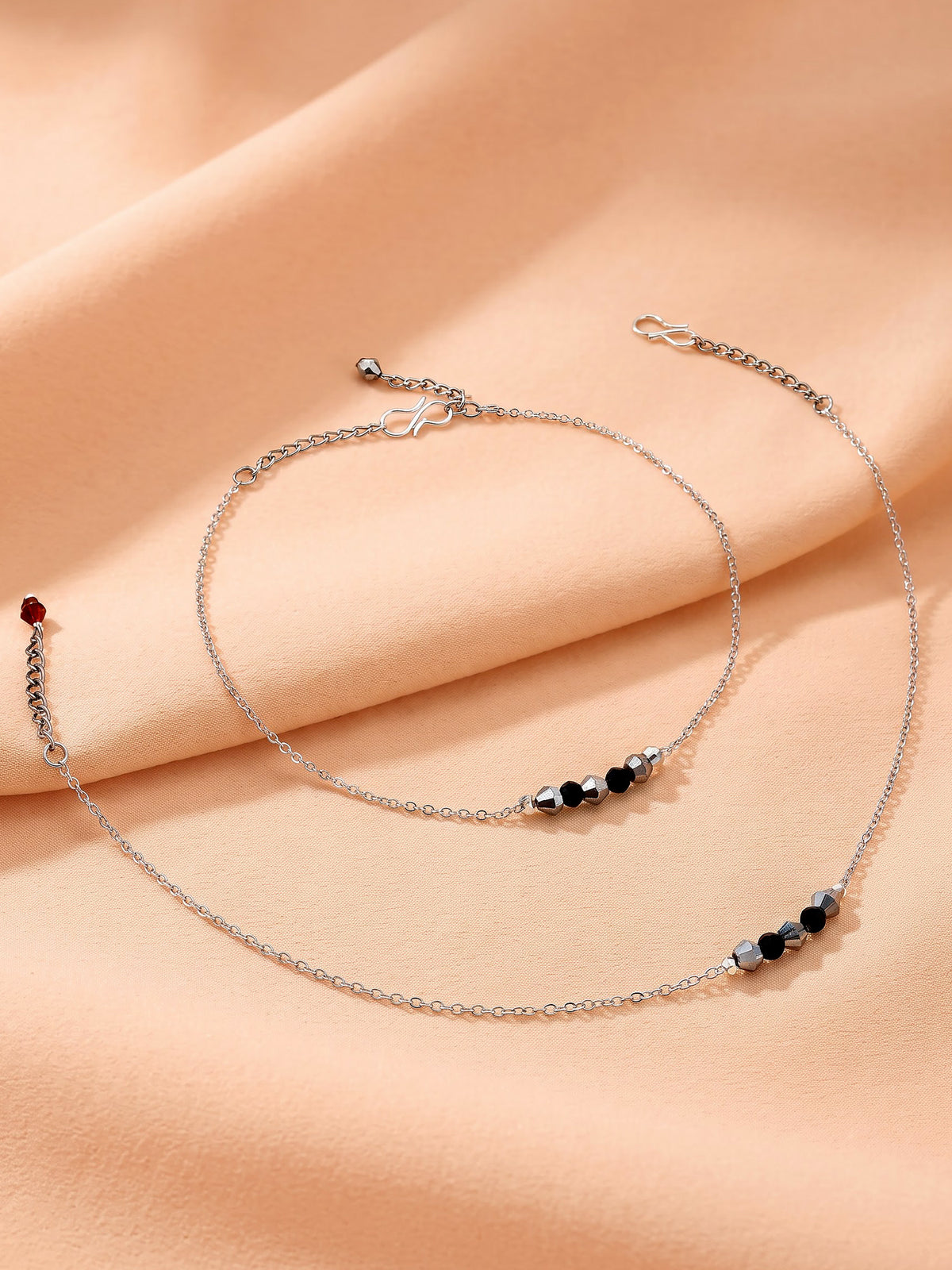Two delicate silver chain anklets with bead accents, one featuring a small red bead and the other a black, silver, and clear bead sequence, displayed on a draped peach fabric background.