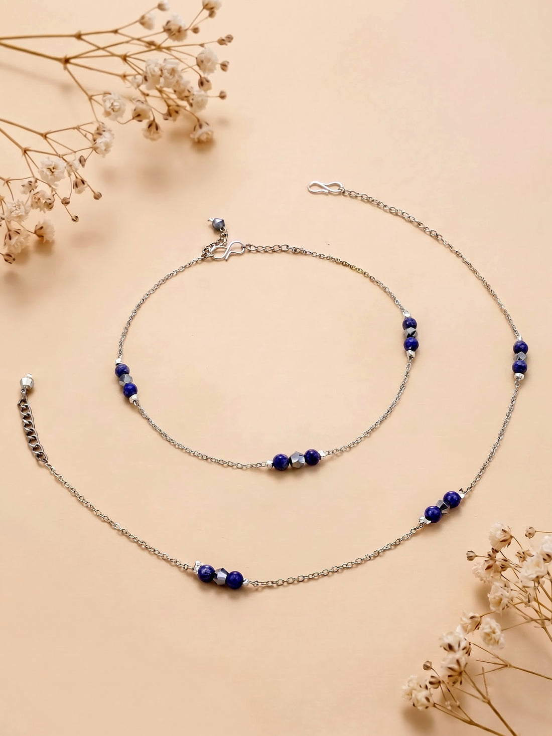 A close-up of a person's feet wearing delicate silver chain anklets with small dark blue beads, shown against a warm beige background.