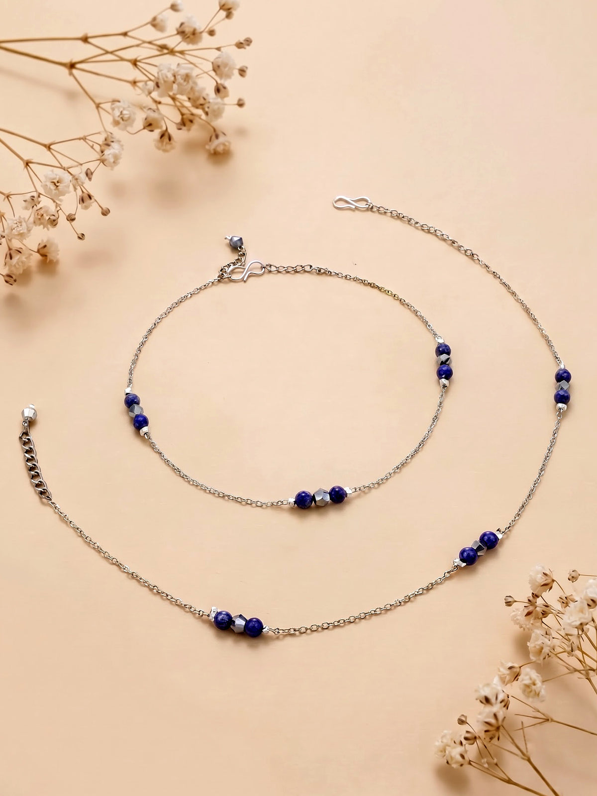 A silver-toned chain anklet with small dark blue beads, displayed on a warm beige background with dried white flowers.