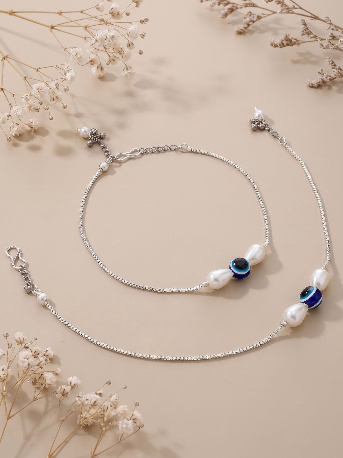 A close-up of a person's feet and lower legs wearing delicate silver anklets with white pearl-like beads and dark blue evil eye accents, set against a warm beige background.