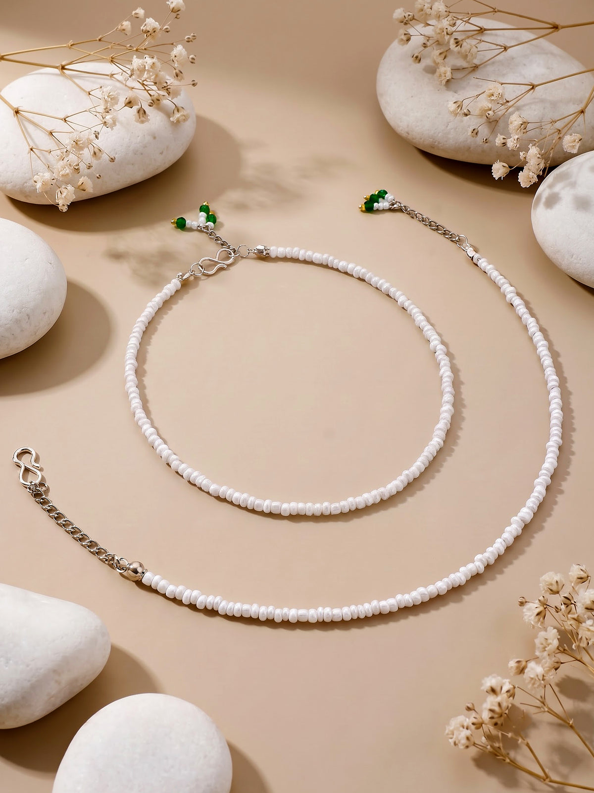 A beaded necklace and matching bracelet laid flat on a warm beige surface with decorative white stones and dried flowers. The jewelry is primarily white beads with silver-toned chain links and small green bead accents near the clasp ends.