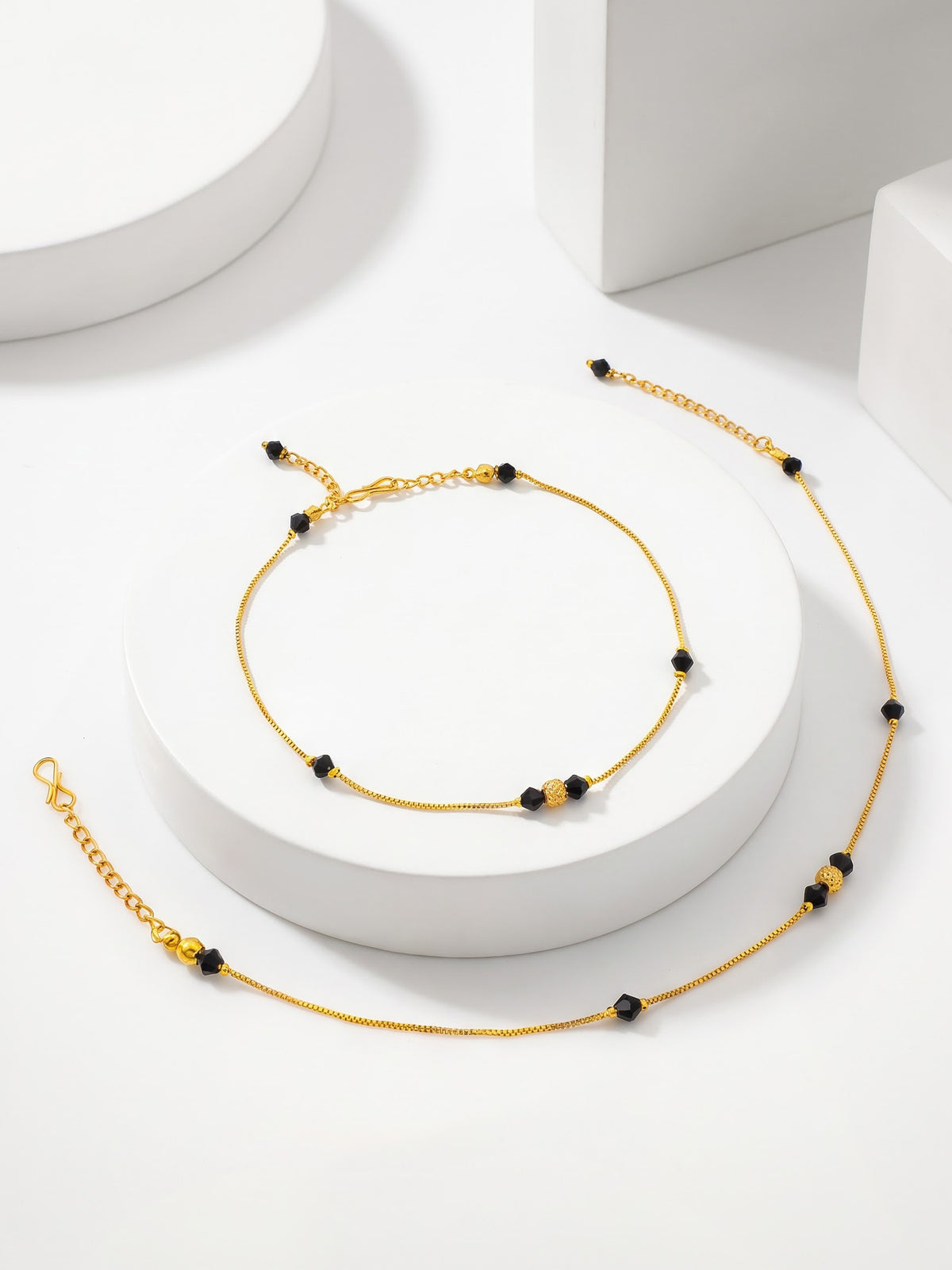 A delicate gold chain anklet with small black beads, displayed on a white cylindrical pedestal against a clean white background.