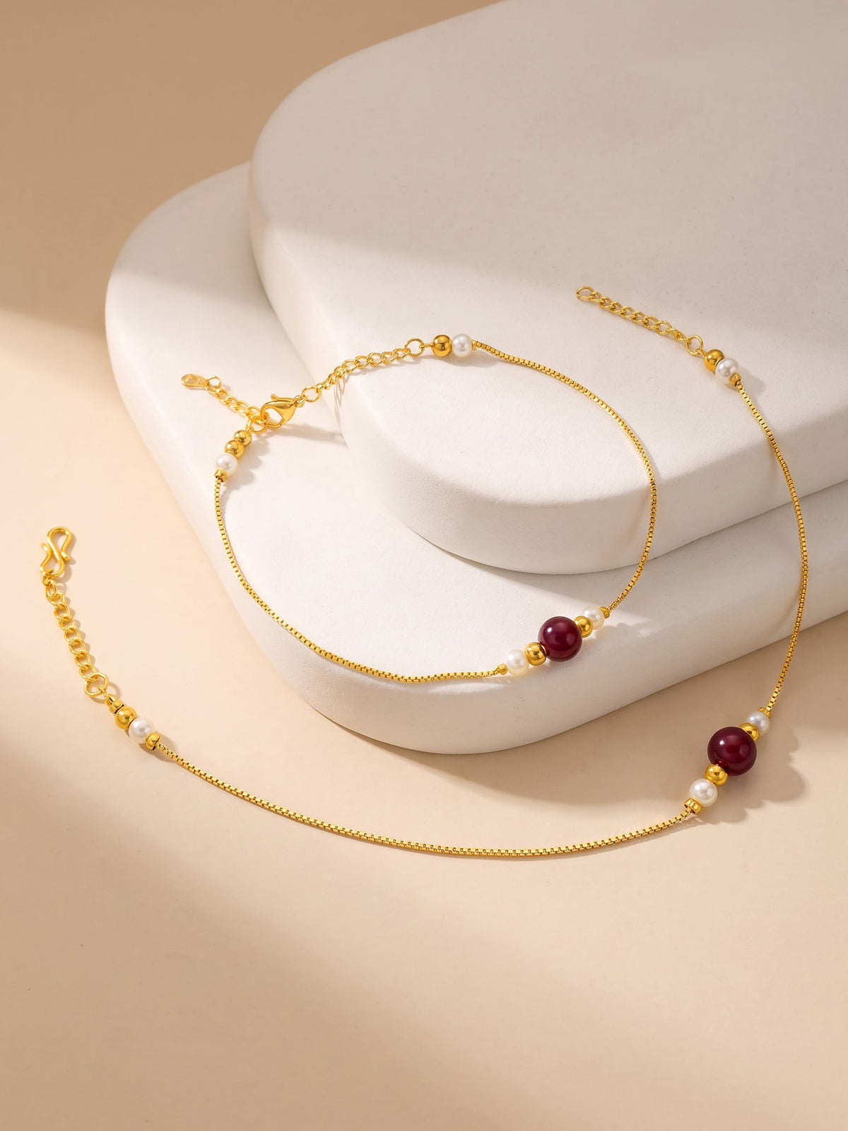 A delicate gold chain anklet with small white pearl-like beads and two dark red round beads, displayed on a white sculptural stand against a warm beige background.