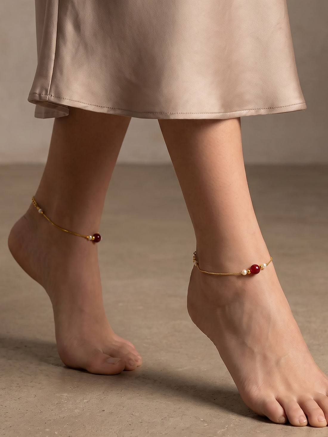 A close-up of a person's feet wearing delicate gold ankle chains decorated with small white pearls and dark red beads, styled with a beige satin skirt.