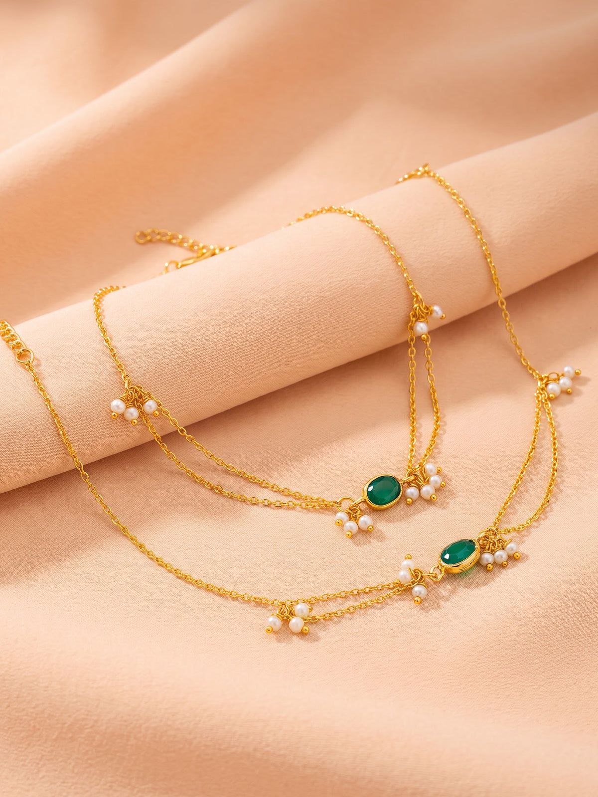 Gold-toned layered necklace with small white pearl clusters and oval green stone accents, displayed on a soft peach fabric background.