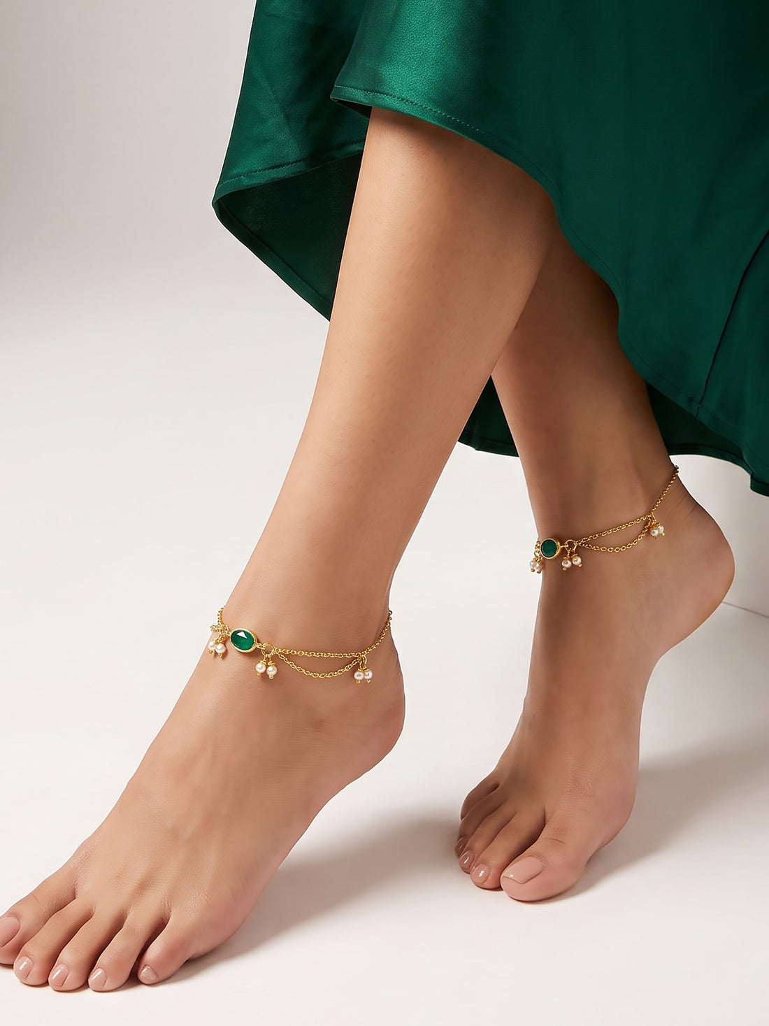 A pair of delicate gold chain anklets with green oval stones and small dangling beads, shown worn on a person's feet with a dark green skirt visible at the top.