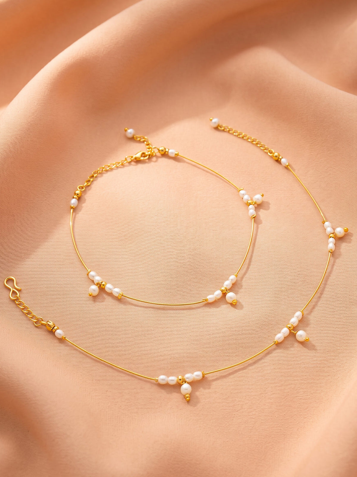 A delicate gold chain anklet decorated with small white freshwater pearls, displayed on a draped peach fabric background.