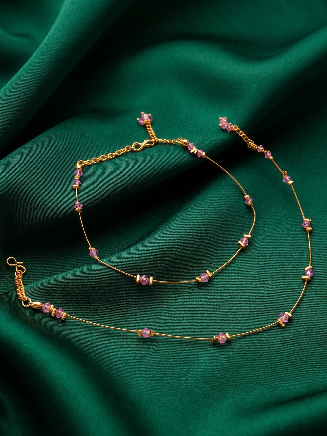 A close-up of a person's bare feet wearing delicate gold anklets with small pink floral details, styled with a green skirt against a warm beige background.