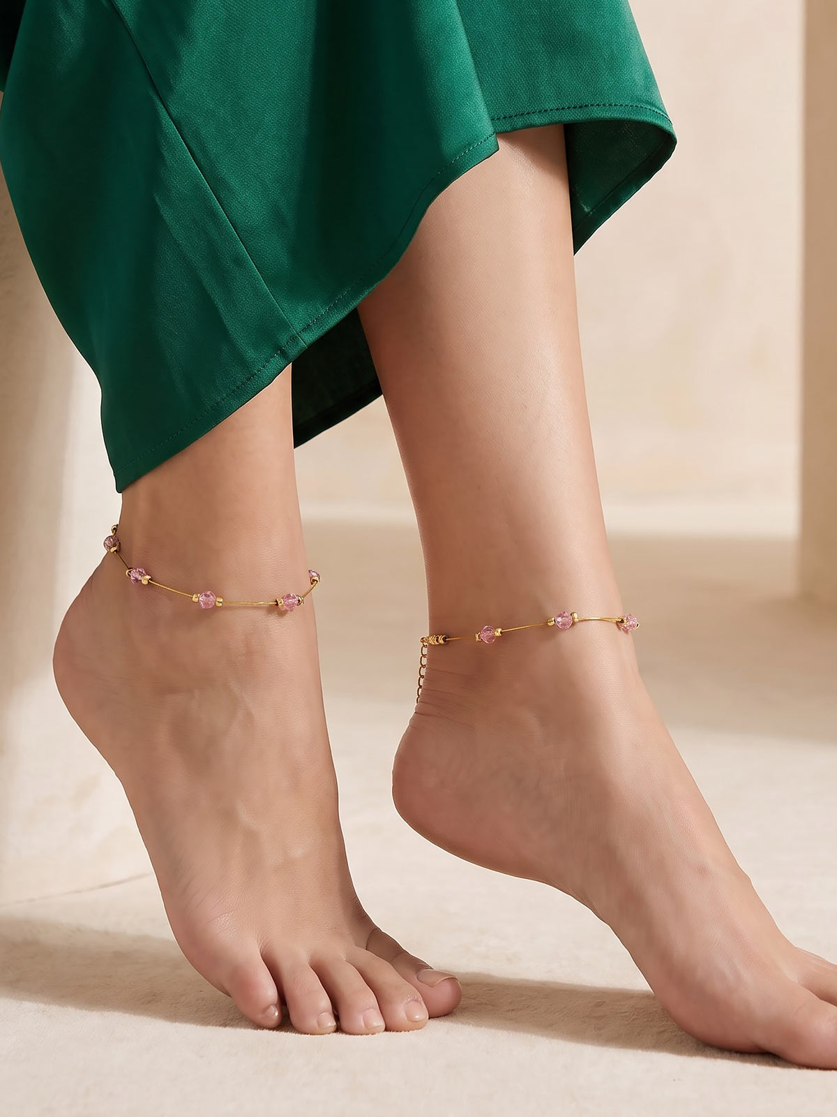 A close-up of a person's bare feet wearing delicate gold anklets with small pink floral details, styled with a green skirt against a warm beige background.