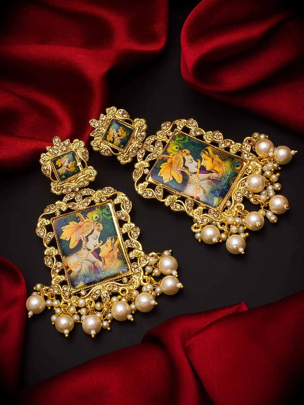 A pair of ornate gold-toned statement earrings with rectangular multicolor enamel portraits and dangling pearl-like beads, arranged on dark fabric with red satin draping.