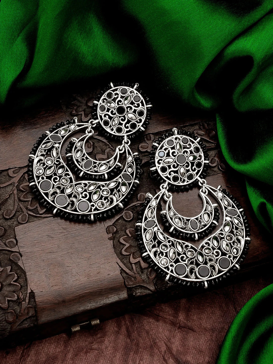 A pair of large silver-toned filigree earrings with circular and crescent-shaped designs, displayed on a carved wooden surface with draped green fabric in the background.