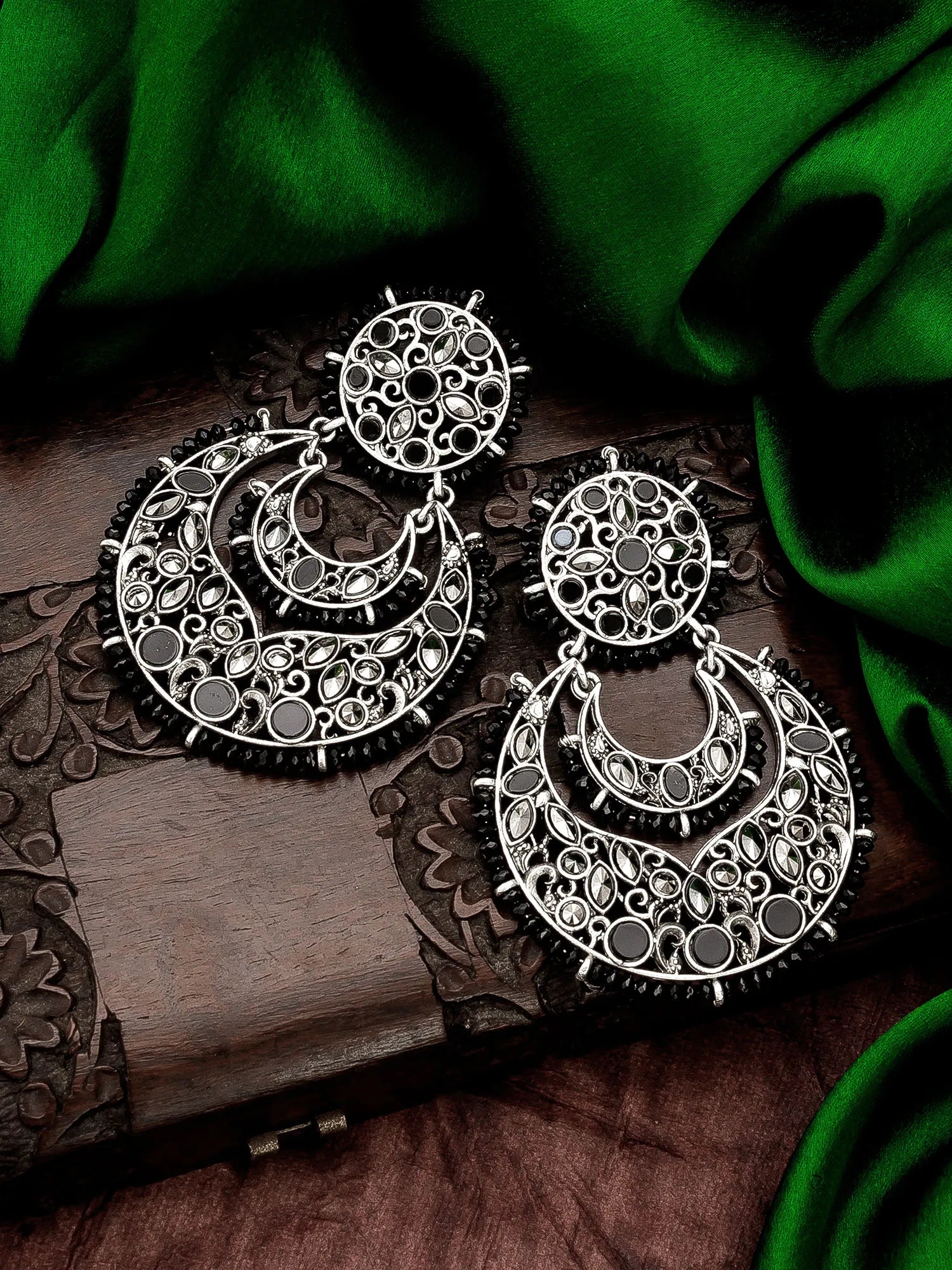 A pair of large silver-toned filigree earrings with circular and crescent-shaped designs, displayed on a carved wooden surface with draped green fabric in the background.