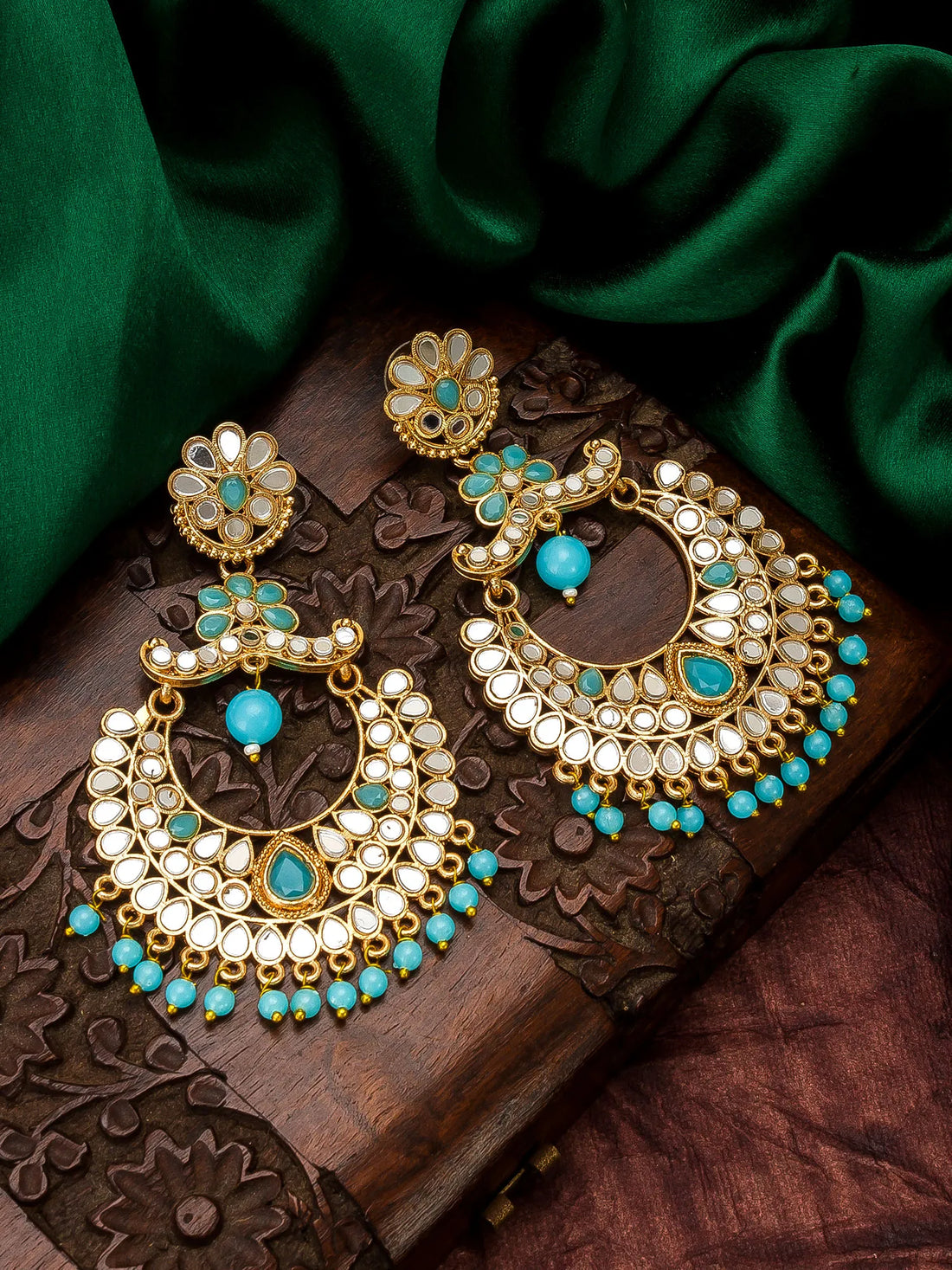 A pair of ornate gold-toned dangle earrings with turquoise blue stones and white inlay, displayed on a carved wooden box with dark green satin fabric in the background.