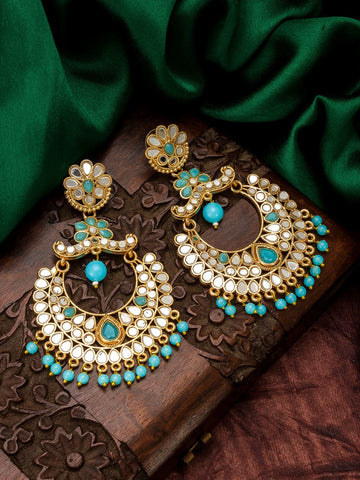 A pair of ornate gold-toned dangle earrings with turquoise blue stones and white inlay, displayed on a carved wooden box with dark green satin fabric in the background.