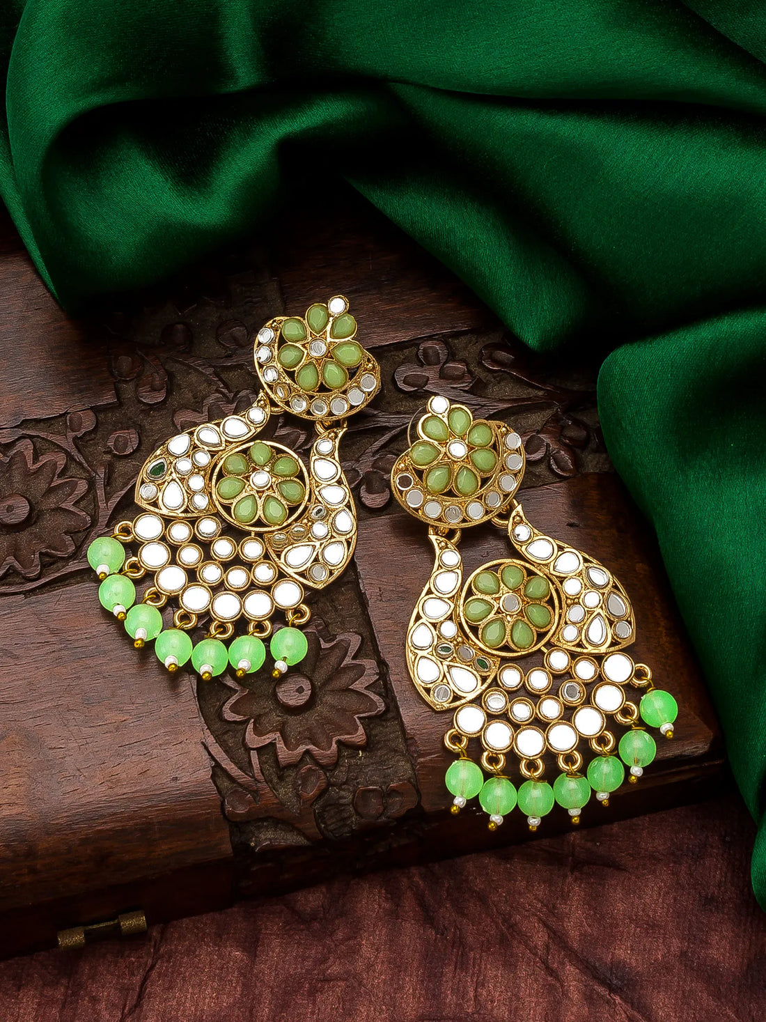 A pair of ornate gold-toned Indian jhumka earrings embellished with green stones and white inlay, displayed on a carved wooden box with draped green fabric in the background.