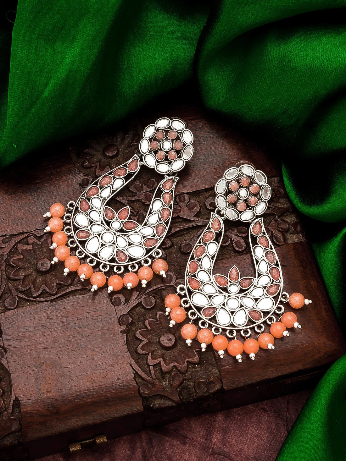 A pair of ornate silver-toned chandelier earrings embellished with white stones, peach-pink teardrop beads, and orange bead fringe, displayed on a carved wooden box with green fabric in the background.