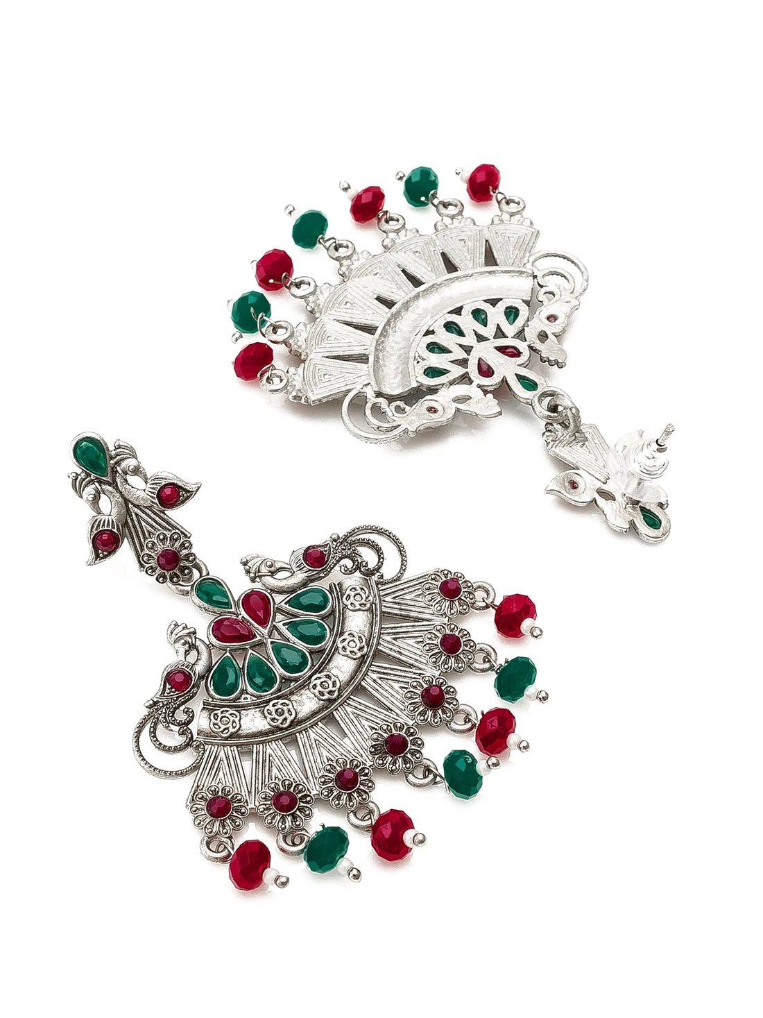 A pair of ornate silver-toned Indian jhumka earrings decorated with green and red stones and dangling bead accents, displayed on a carved dark wooden box with green fabric in the background.