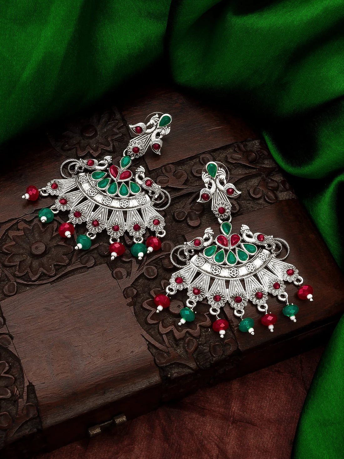 A pair of ornate silver-toned Indian jhumka earrings decorated with green and red stones and dangling bead accents, displayed on a carved dark wooden box with green fabric in the background.