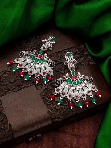 A pair of ornate silver-toned Indian jhumka earrings decorated with green and red stones and dangling bead accents, displayed on a carved dark wooden box with green fabric in the background.