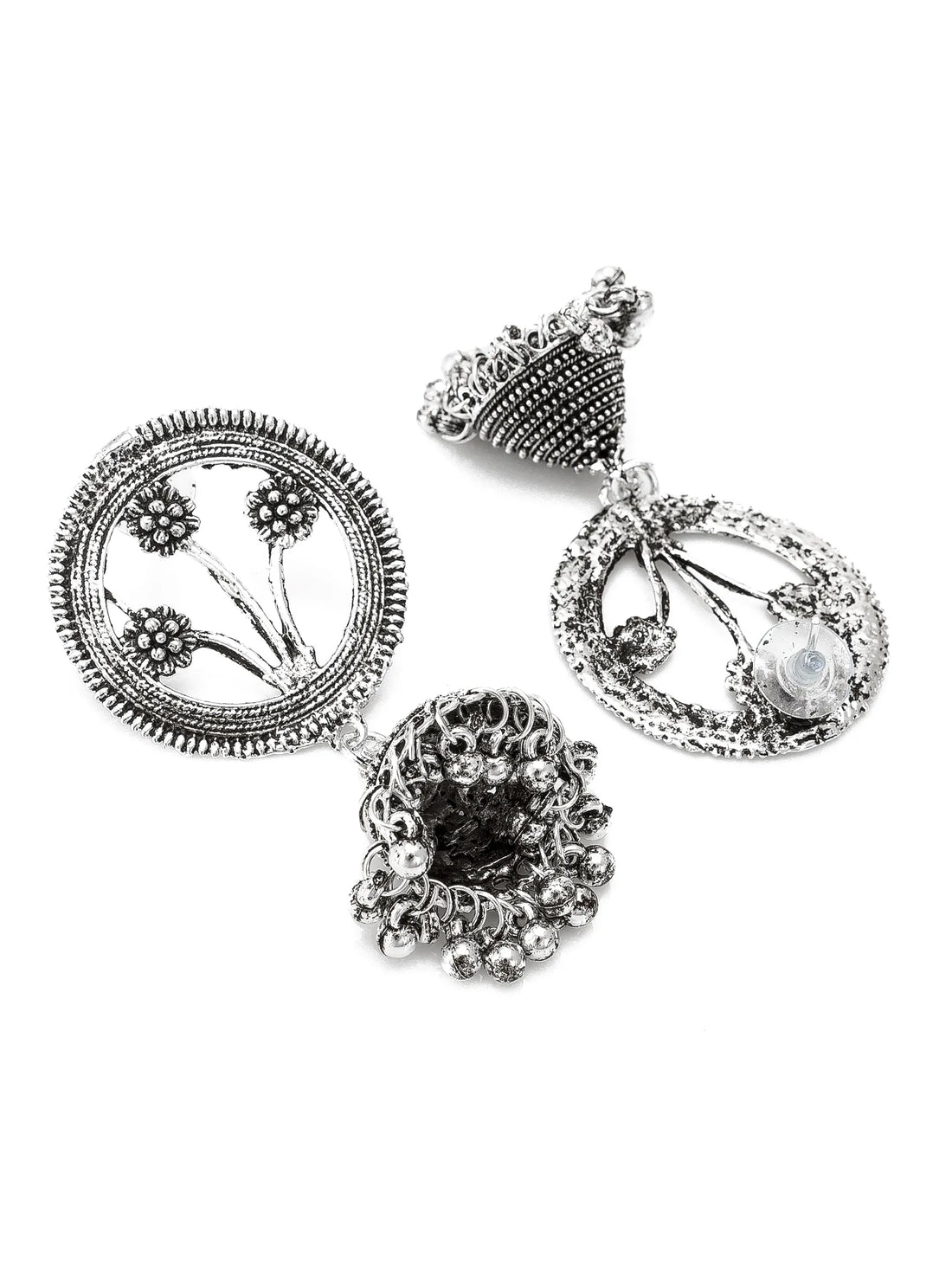 A pair of silver-toned Indian Jhumka earrings with intricate floral motifs and dangling bell-shaped charms, displayed on a carved wooden block with draped green fabric in the background.