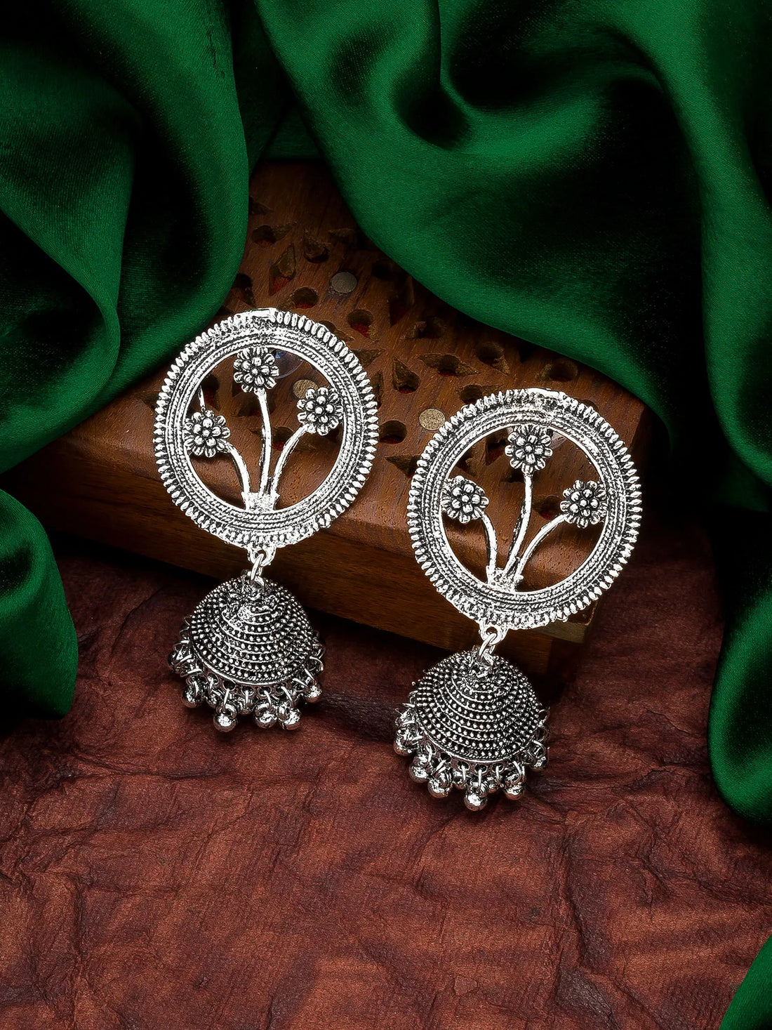 A pair of silver-toned Indian Jhumka earrings with intricate floral motifs and dangling bell-shaped charms, displayed on a carved wooden block with draped green fabric in the background.