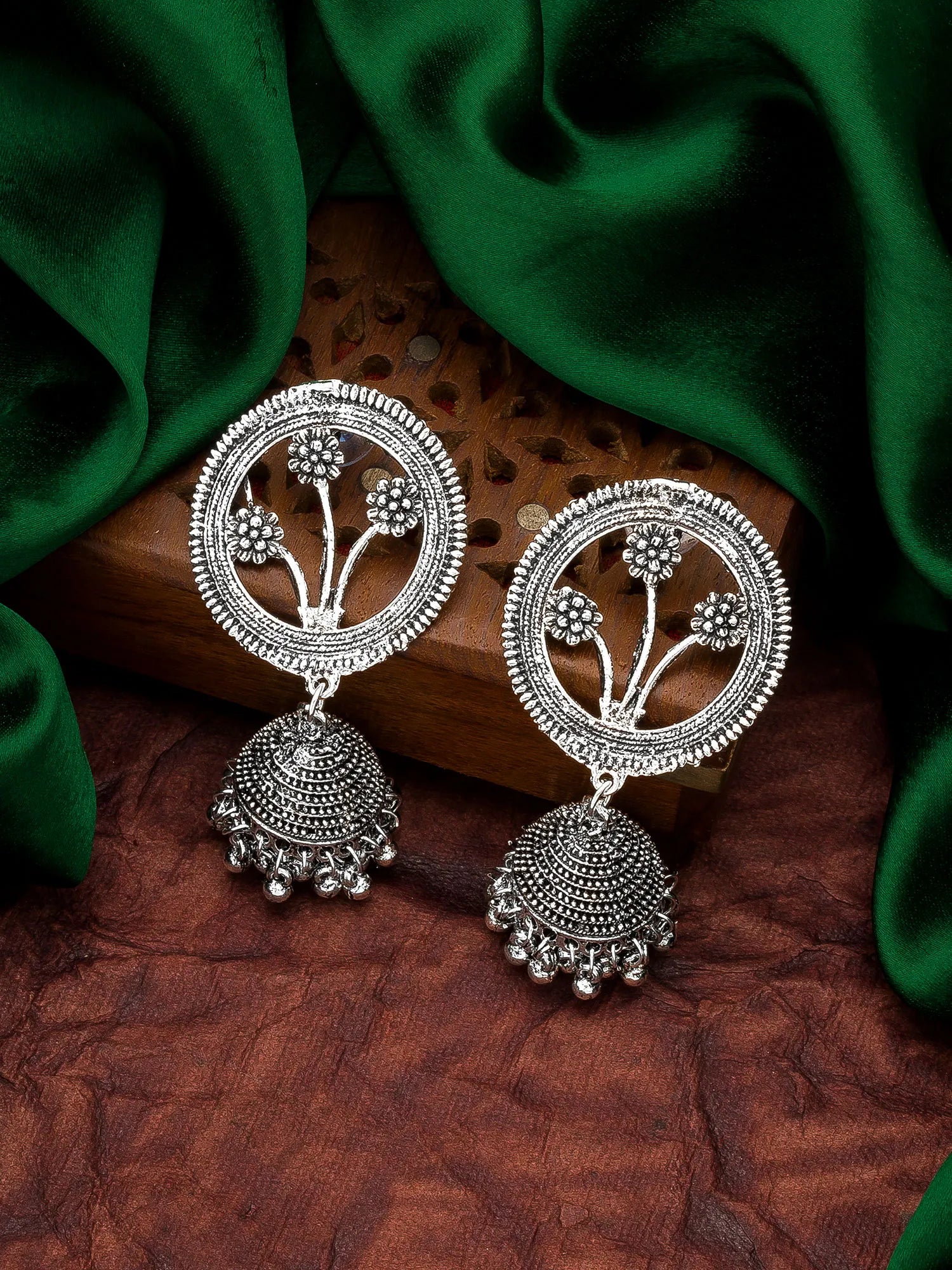 A pair of silver-toned Indian Jhumka earrings with intricate floral motifs and dangling bell-shaped charms, displayed on a carved wooden block with draped green fabric in the background.