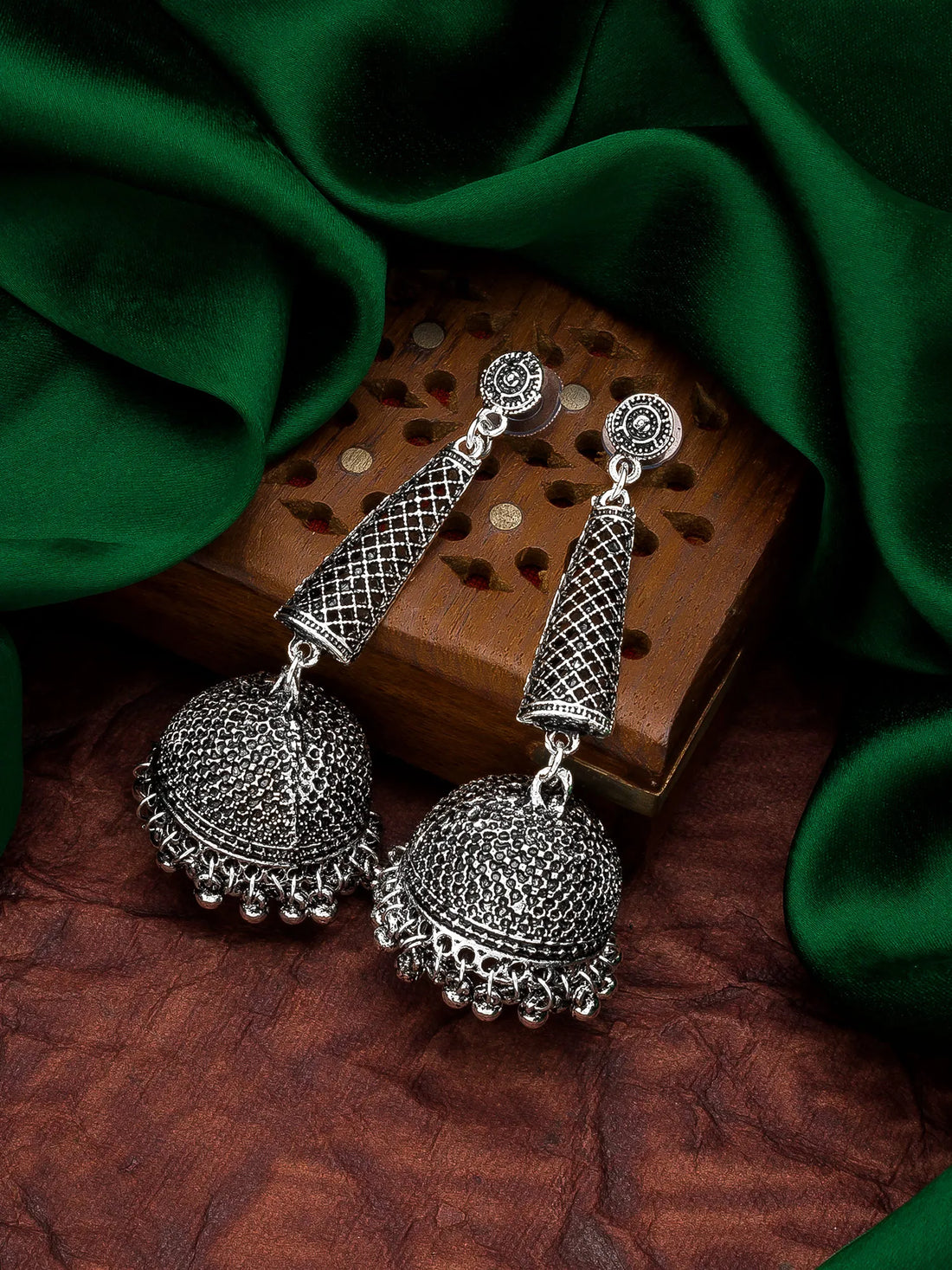 A pair of silver-toned Indian jhumka earrings with intricate filigree detailing, displayed on a wooden block and draped with dark green silk fabric.