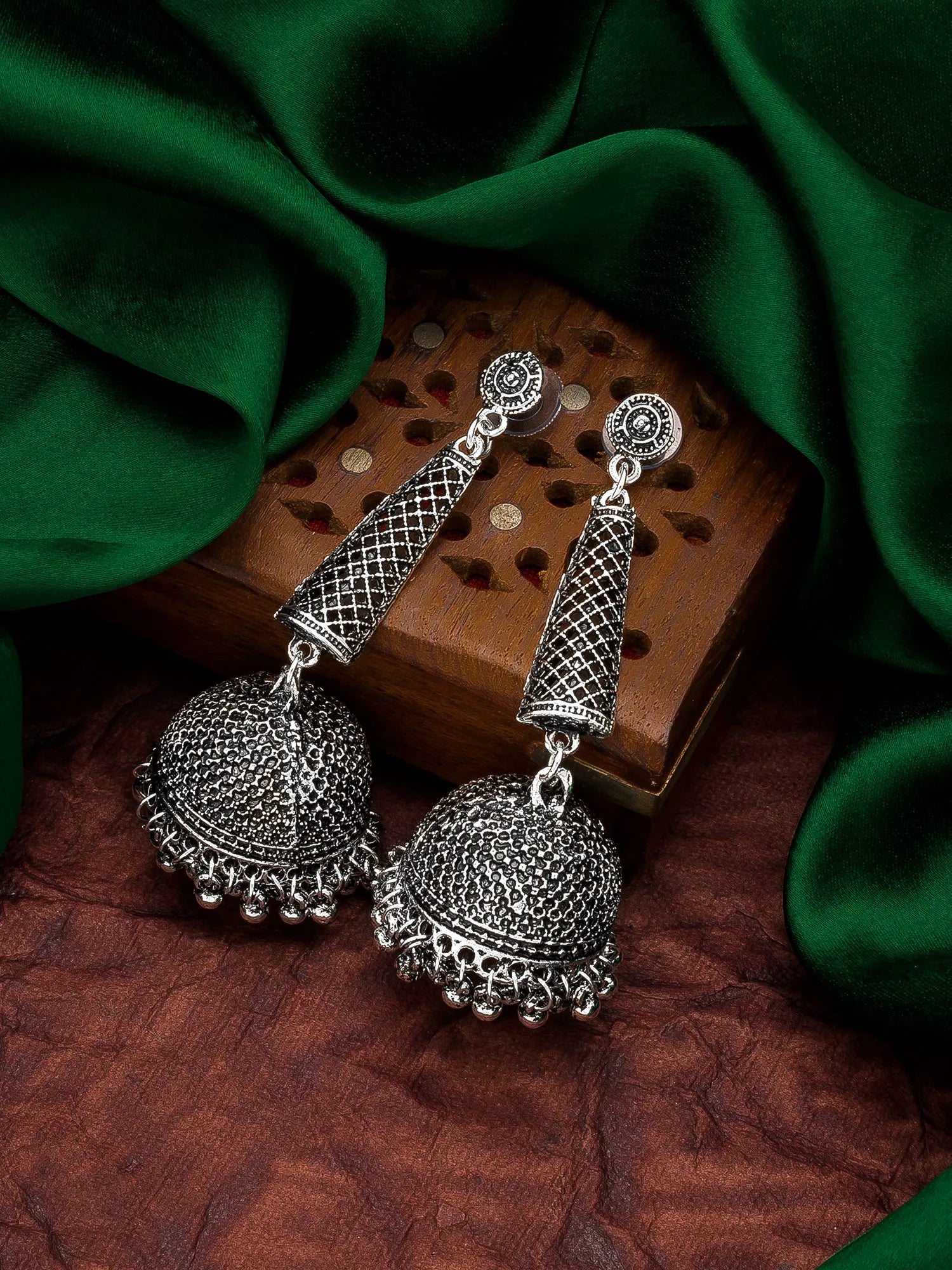 A pair of silver-toned Indian jhumka earrings with intricate filigree detailing, displayed on a wooden block and draped with dark green silk fabric.