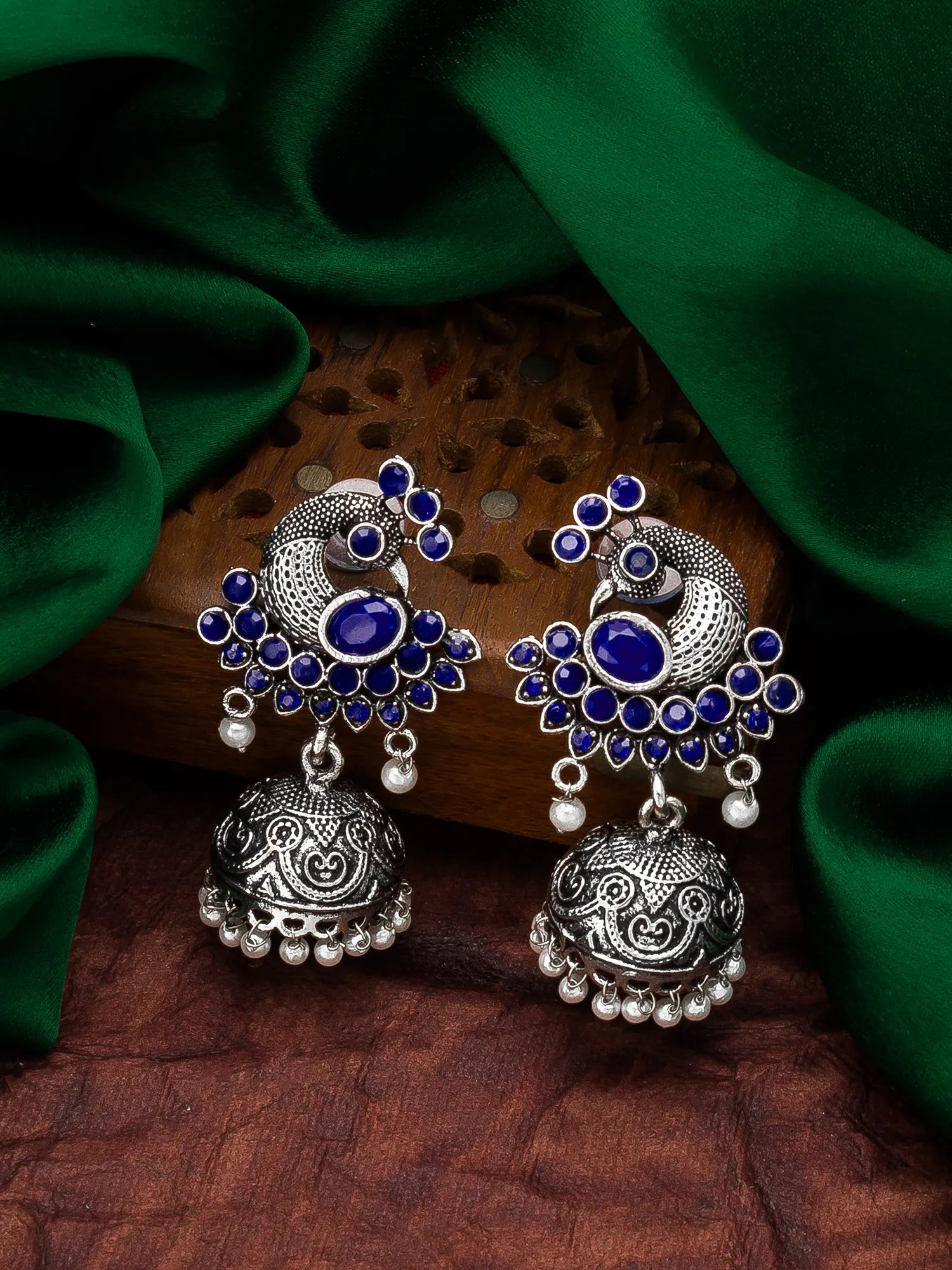 A pair of traditional Indian silver-toned jhumka earrings with deep blue stones and small white bead accents, displayed on a carved wooden block with draped green fabric in the background.