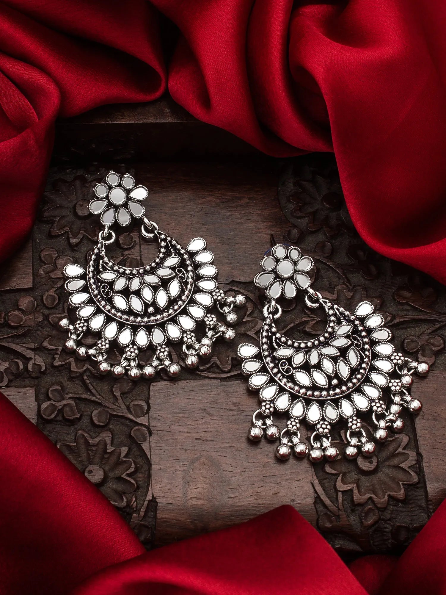 A pair of ornate silver-toned Indian jhumka earrings with floral studs and teardrop-shaped dangling elements, displayed on a carved dark wooden surface surrounded by draped red satin fabric.