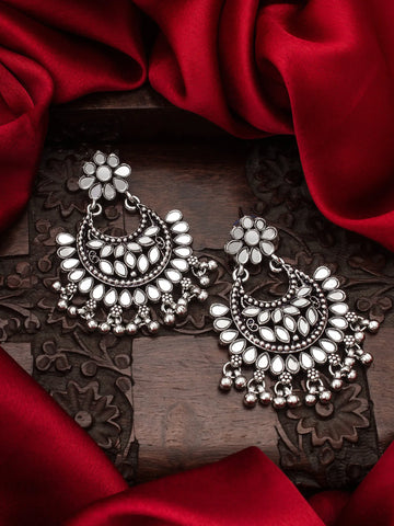A pair of ornate silver-toned Indian jhumka earrings with floral studs and teardrop-shaped dangling elements, displayed on a carved dark wooden surface surrounded by draped red satin fabric.