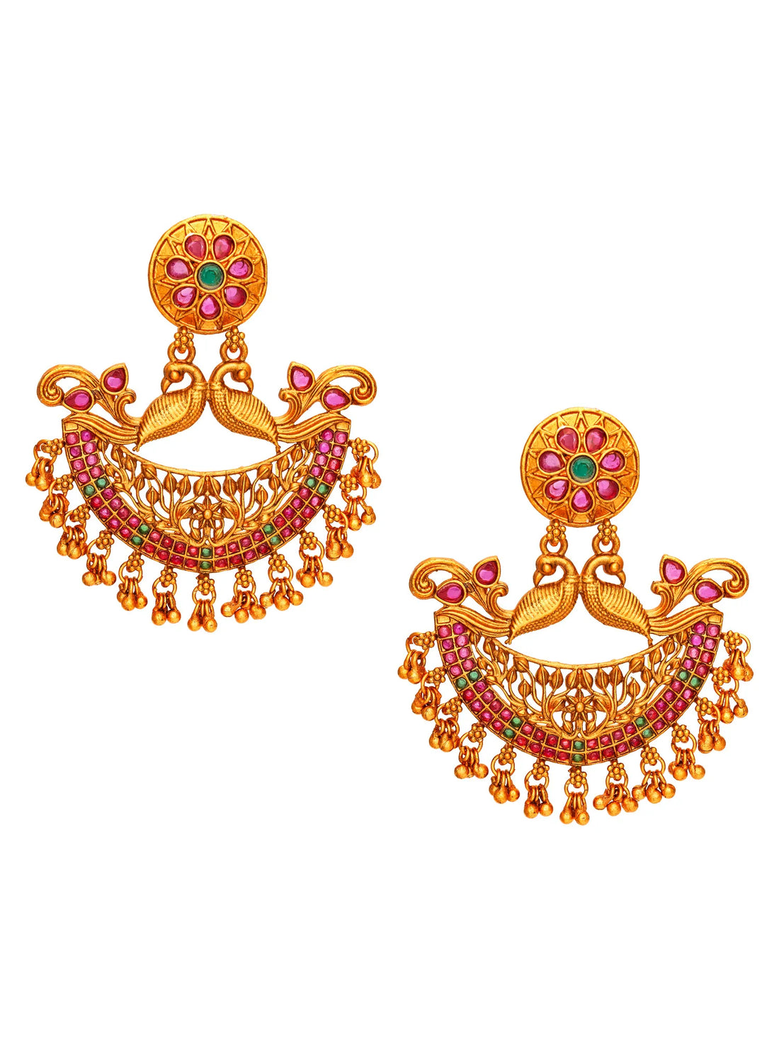 A pair of ornate gold-toned traditional Indian earrings with peacock motifs, red and green stone embellishments, and dangling bead chains, arranged on a dark wood surface with draped red fabric.