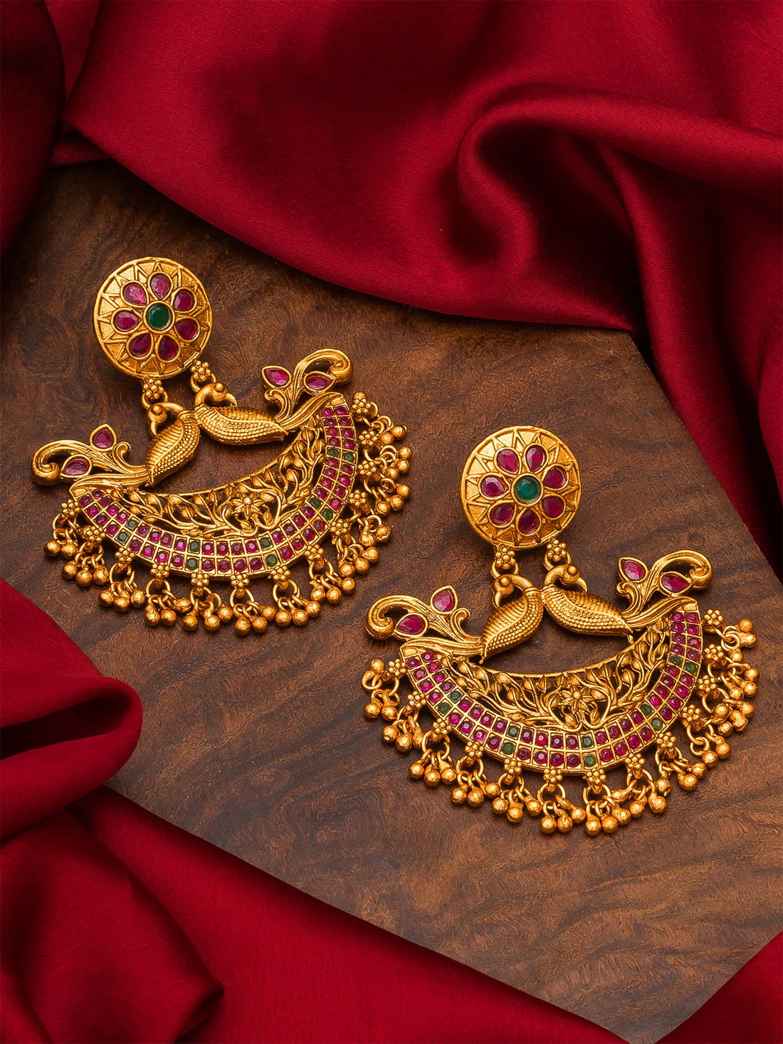A pair of ornate gold-toned traditional Indian earrings with peacock motifs, red and green stone embellishments, and dangling bead chains, arranged on a dark wood surface with draped red fabric.
