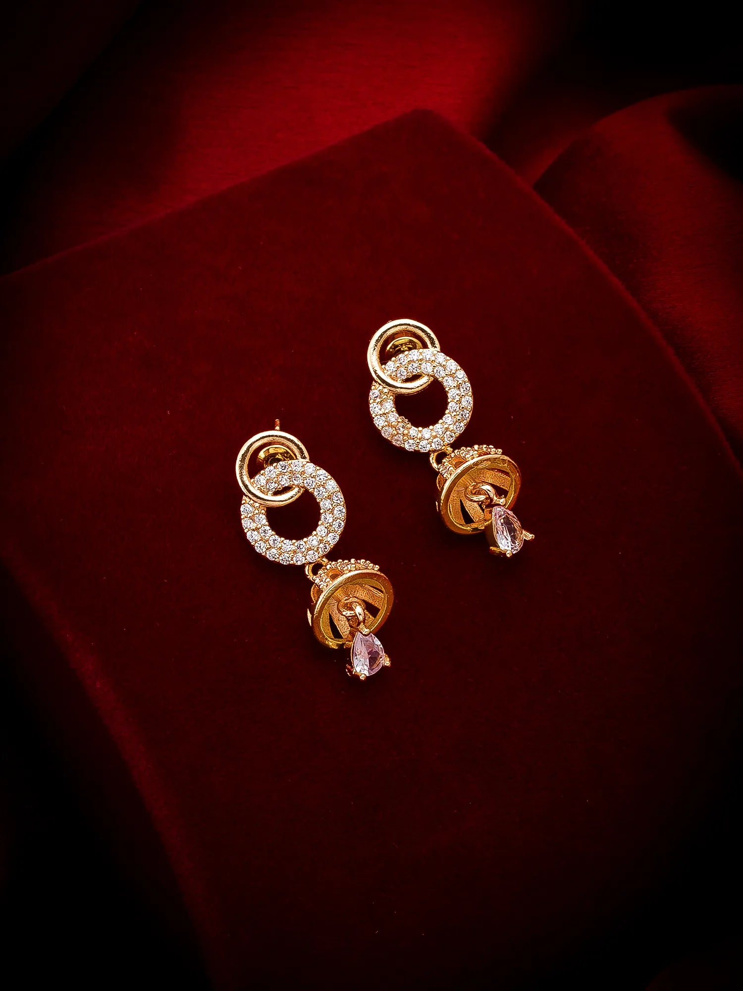 A pair of gold-toned statement earrings with pavé-set clear stones in circular openwork designs, each hanging from a gold ring and finished with a dangling faceted crystal, displayed on a deep red velvet surface.