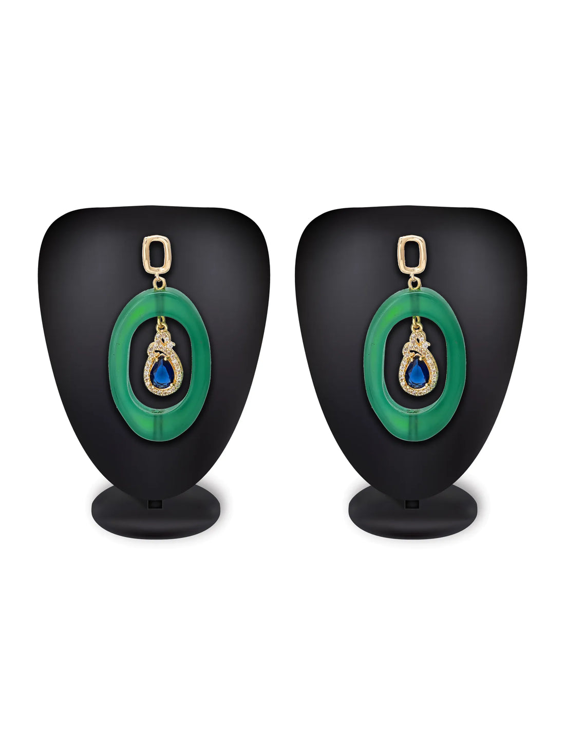 A pair of green oval drop earrings with gold-tone hardware and a teardrop-shaped dark blue stone in the center, displayed on a bright turquoise background with decorative pink flowers and a white plate edge.