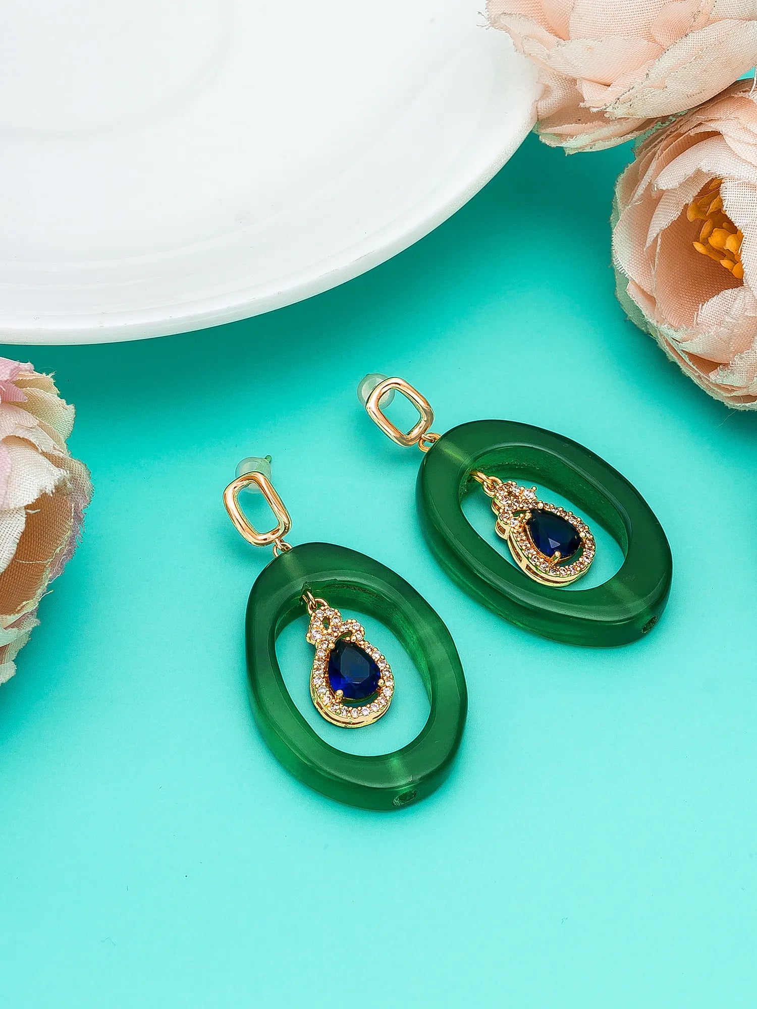 A pair of green oval drop earrings with gold-tone hardware and a teardrop-shaped dark blue stone in the center, displayed on a bright turquoise background with decorative pink flowers and a white plate edge.