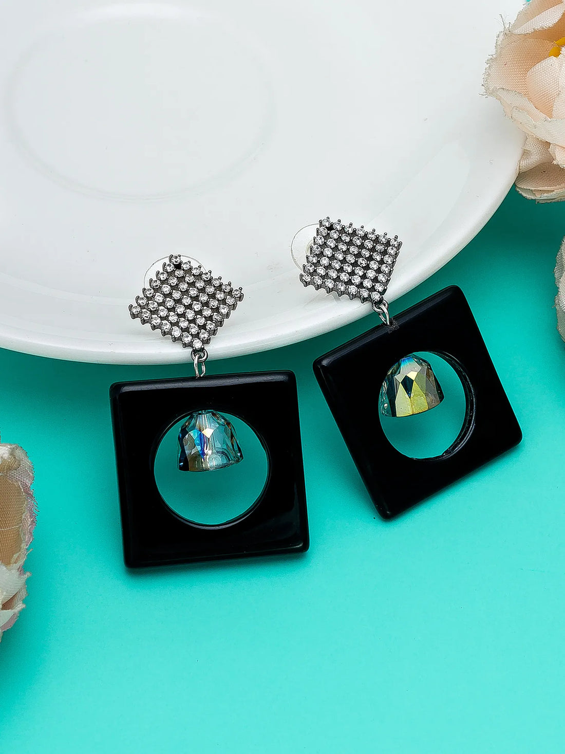 A pair of black square drop earrings with silver rhinestone-embellished posts and a faceted iridescent gemstone in the center, displayed on a white plate over a turquoise background.