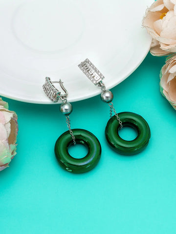 A pair of drop earrings with silver-toned huggie hoops, silver beaded chains, and glossy dark green circular pendant charms, styled on a white plate over a bright turquoise background with soft pink flowers.