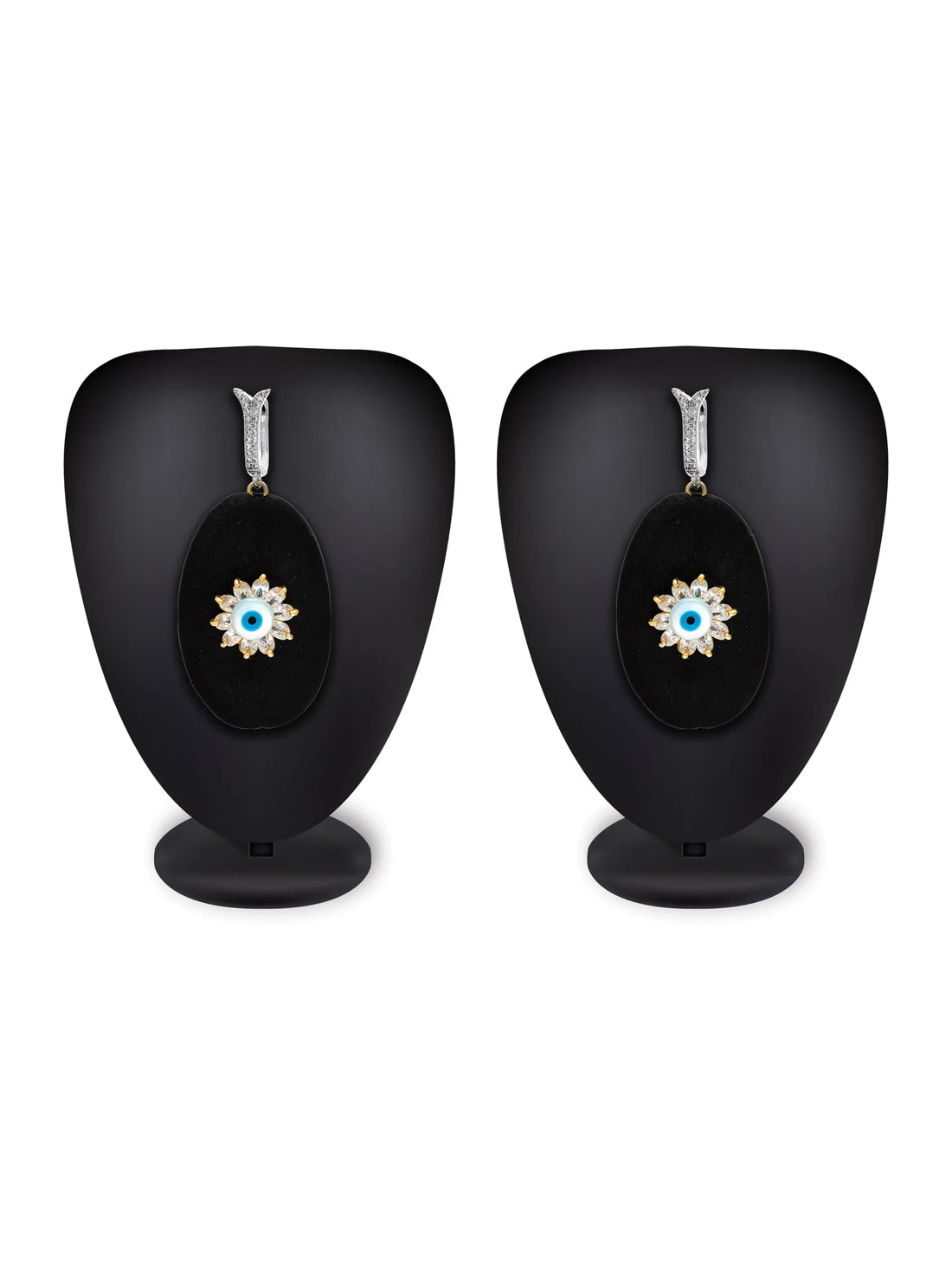 A pair of black oval drop earrings with silver-toned ear clips and gold-toned evil eye charms, displayed on a turquoise background with a white plate and peach flowers.