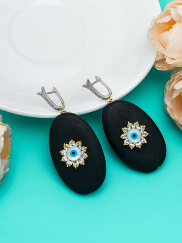 A pair of black oval drop earrings with silver-toned ear clips and gold-toned evil eye charms, displayed on a turquoise background with a white plate and peach flowers.