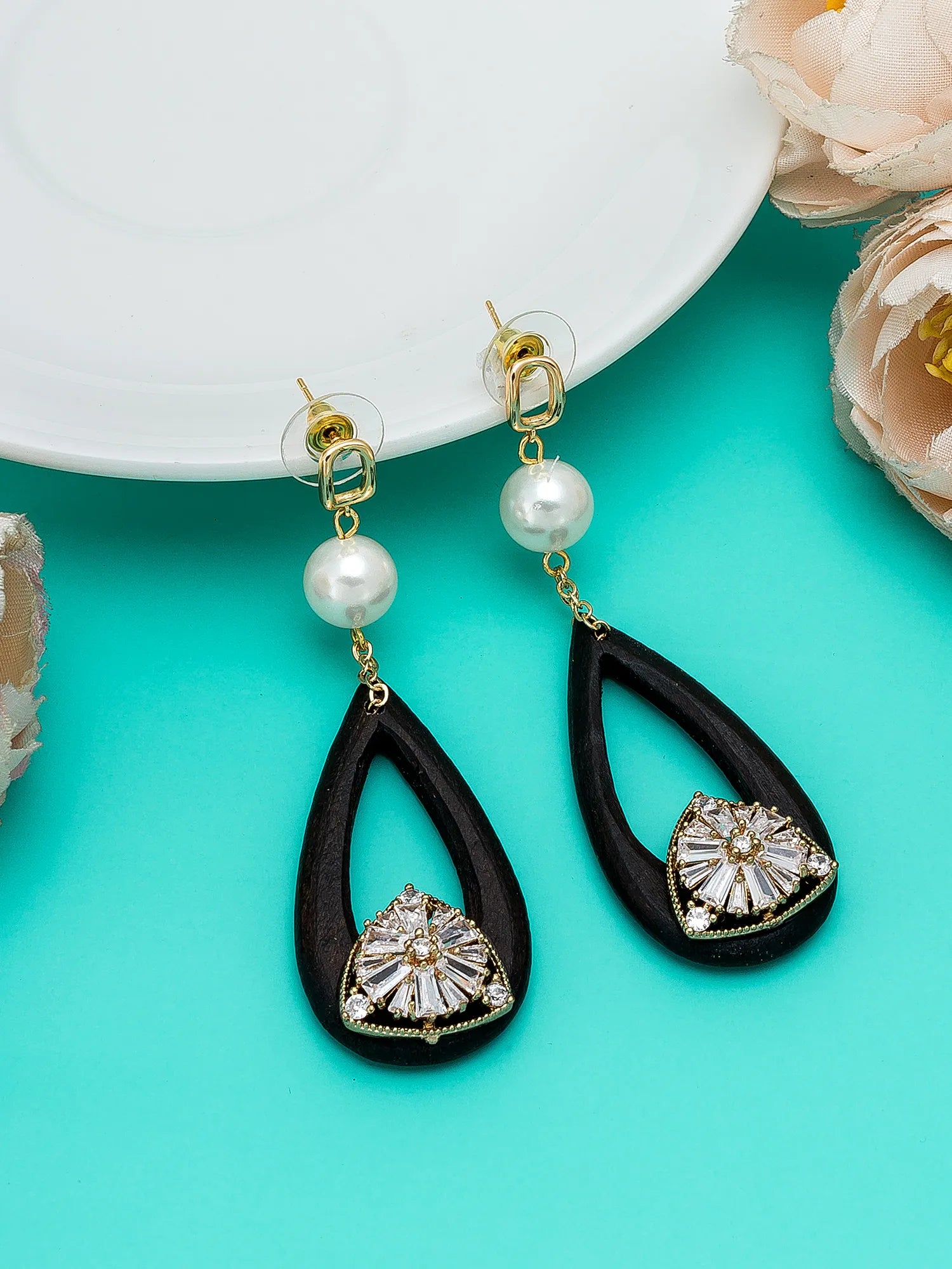 A pair of black teardrop-shaped drop earrings with gold-tone posts, white pearl beads, and a clear rhinestone floral design at the bottom, displayed on a turquoise background with a white plate and pale flowers.