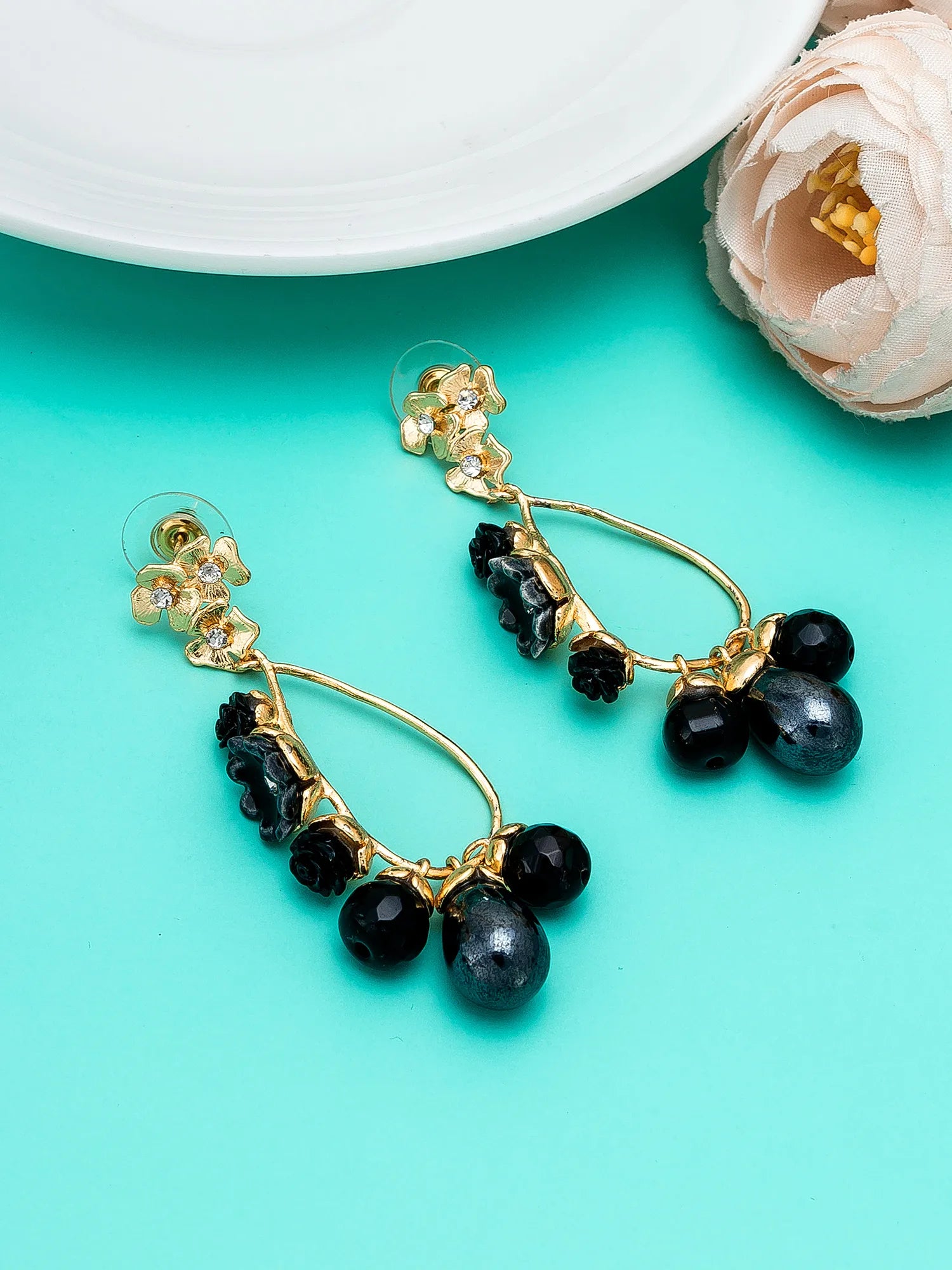 Gold-tone drop earrings with black faceted beads and crystal accents, displayed on a bright turquoise background with decorative floral props.