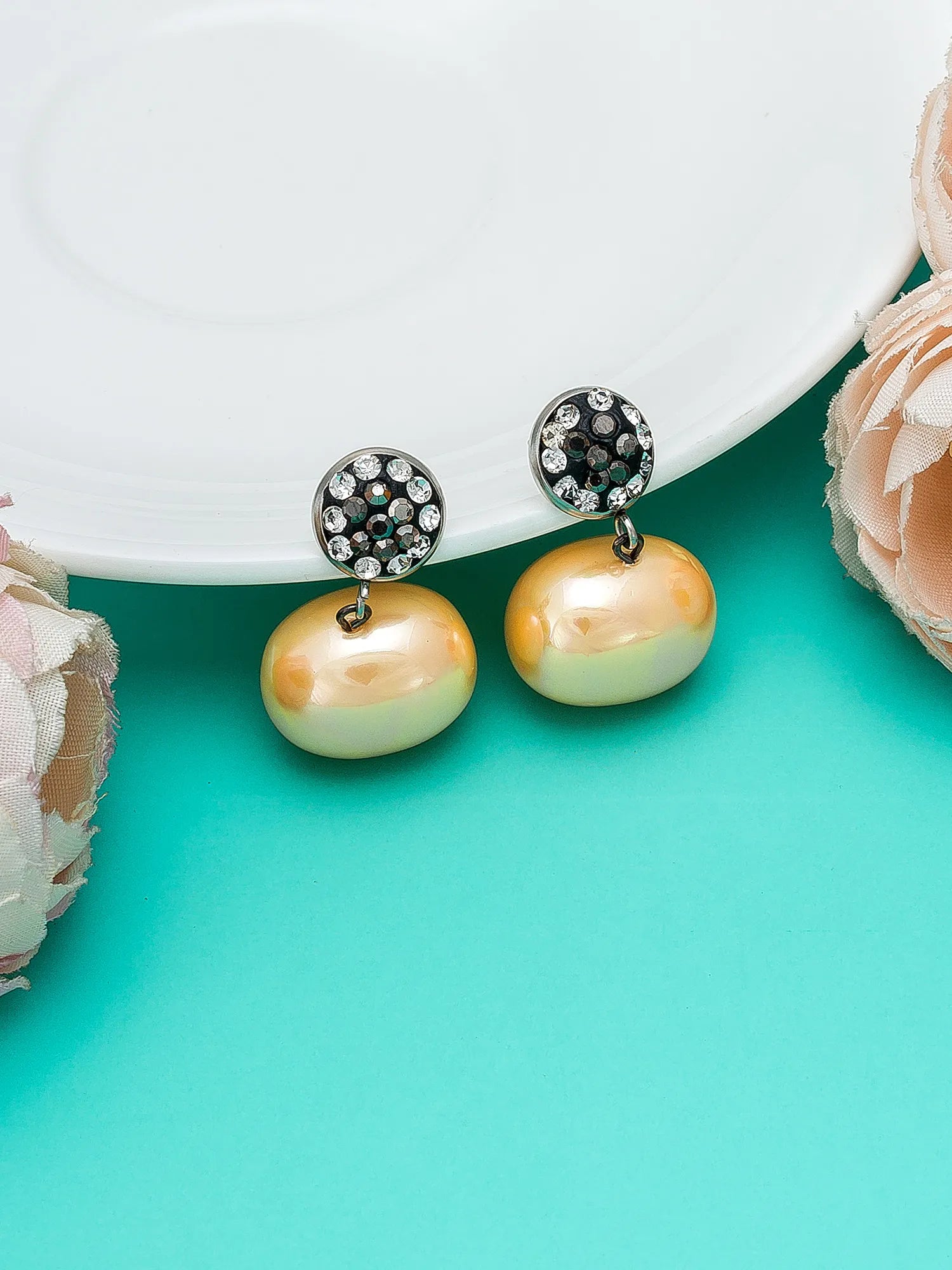 A pair of drop earrings featuring large, lustrous pearlescent beads in a soft peach and pale yellow-green gradient, suspended from silver-toned studs adorned with small clear crystals, styled on a turquoise background with a white plate and pink flowers.