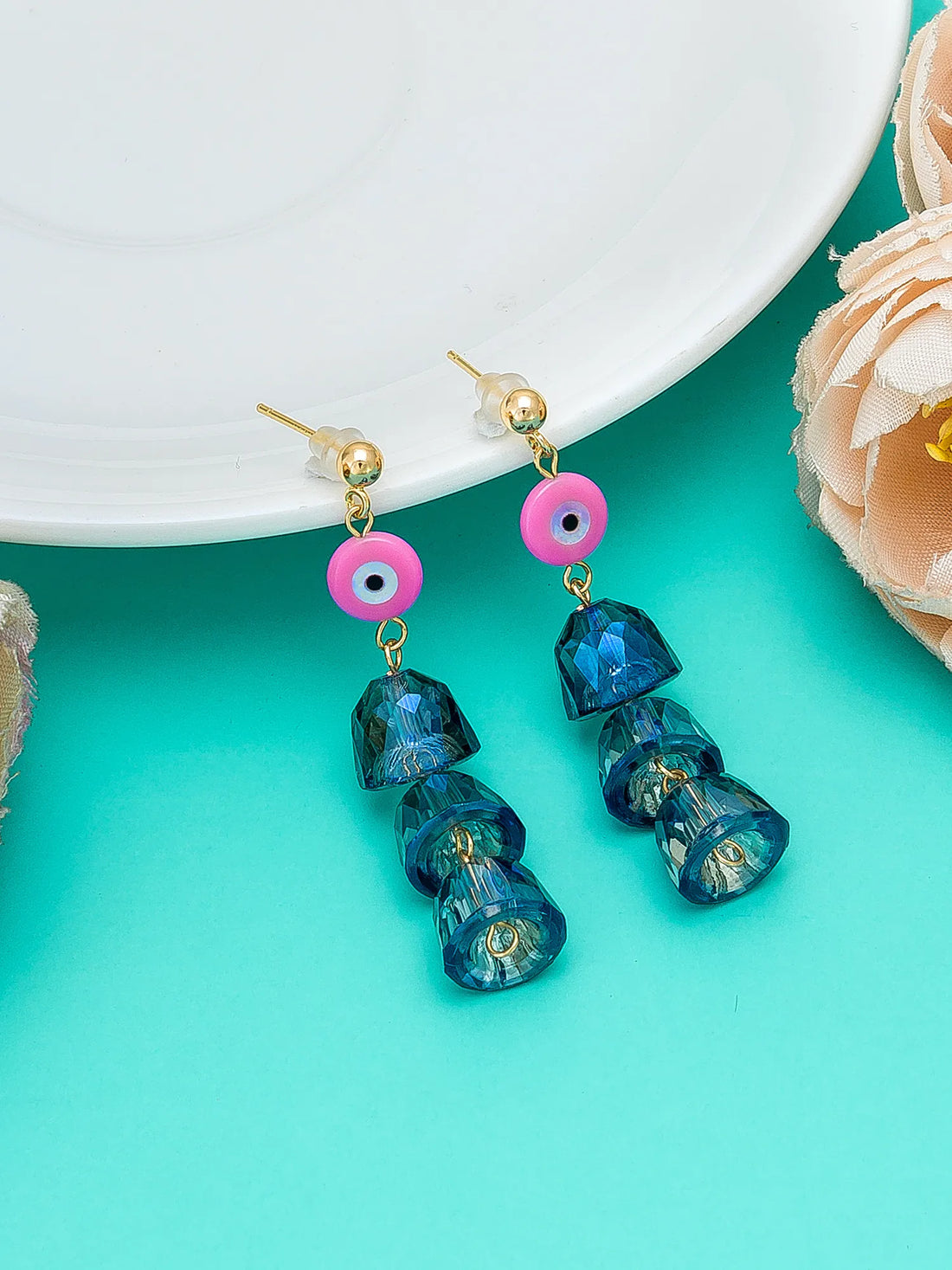 A pair of dangling earrings with gold-tone posts, pink evil eye charms, and three faceted dark blue beads, displayed on a turquoise background with a white plate and a peach flower prop.