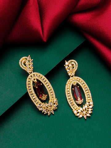 A pair of ornate gold-toned dangle earrings with marquise-cut deep red gemstones and pavé-set clear stones, displayed on a dark green background with draped red satin fabric.
