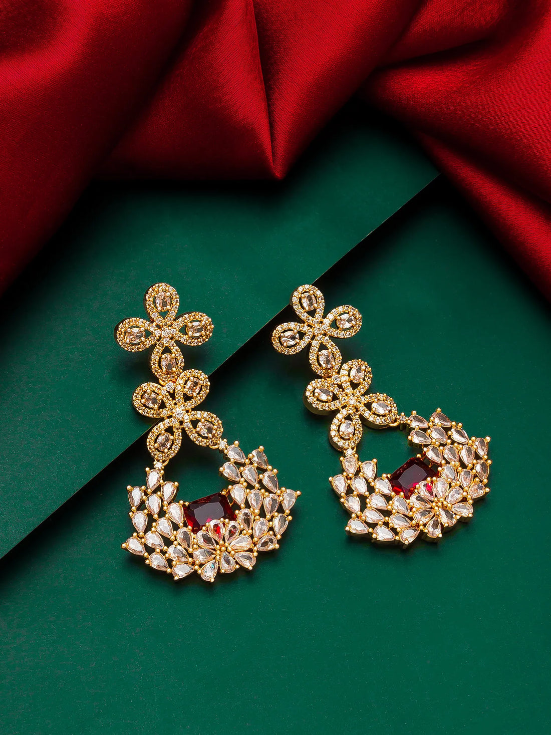 A pair of ornate gold-toned chandelier earrings with clear stones and a central deep red stone, displayed on a dark green background with red satin fabric.