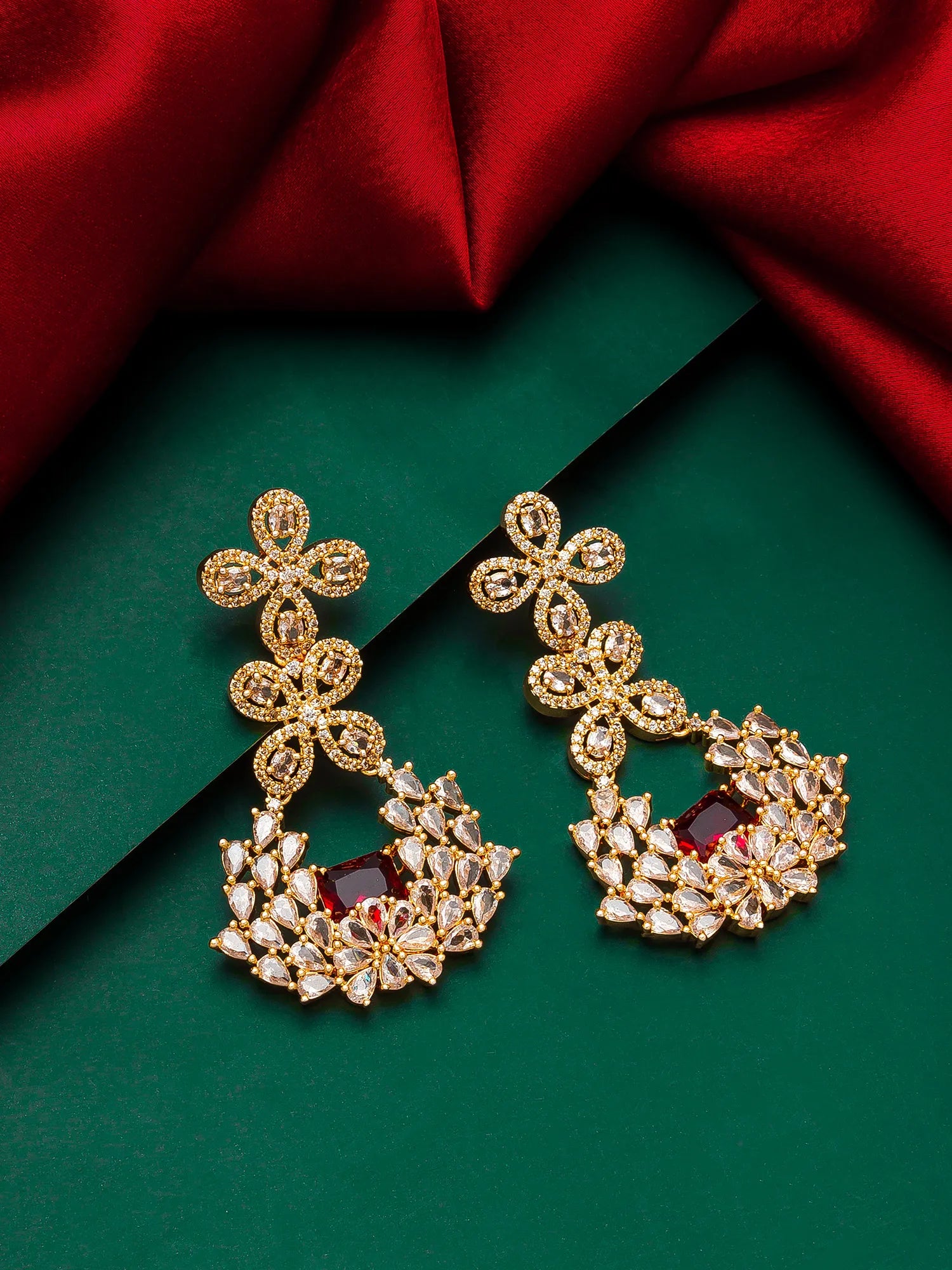 A pair of ornate gold-toned chandelier earrings with clear stones and a central deep red stone, displayed on a dark green background with red satin fabric.
