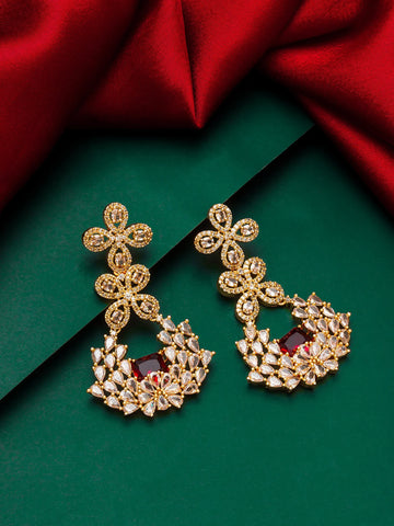 A pair of ornate gold-toned chandelier earrings with clear stones and a central deep red stone, displayed on a dark green background with red satin fabric.