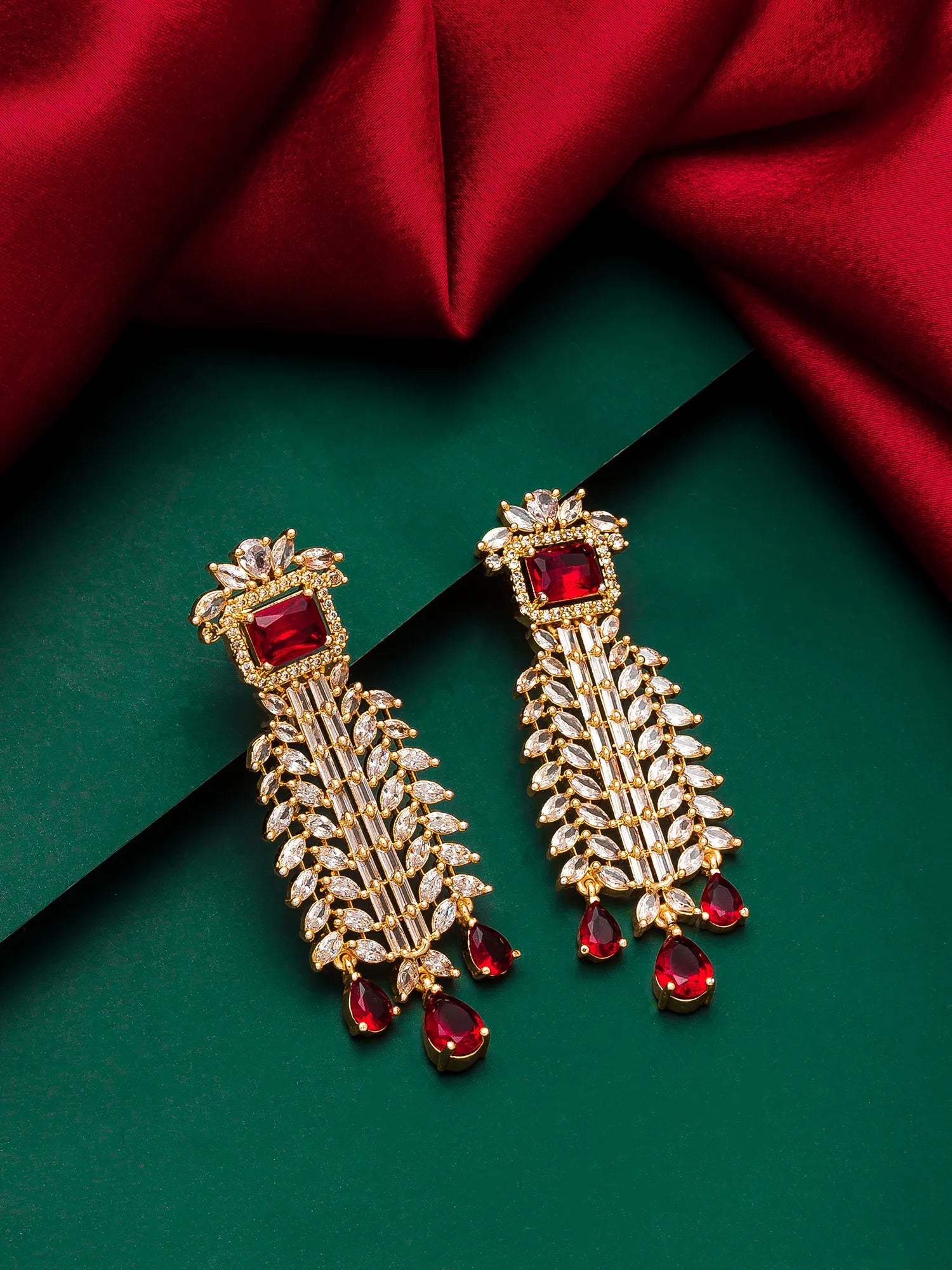 A pair of gold-toned traditional Indian chandelier earrings with rectangular red gemstones and teardrop red drops, embellished with small clear stones, displayed on a dark green background with a draped red fabric.