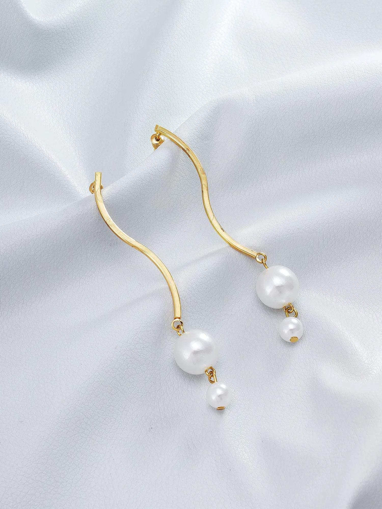 Gold-tone wavy hoop earrings with dangling white pearl drops, displayed on a soft white satin fabric background.